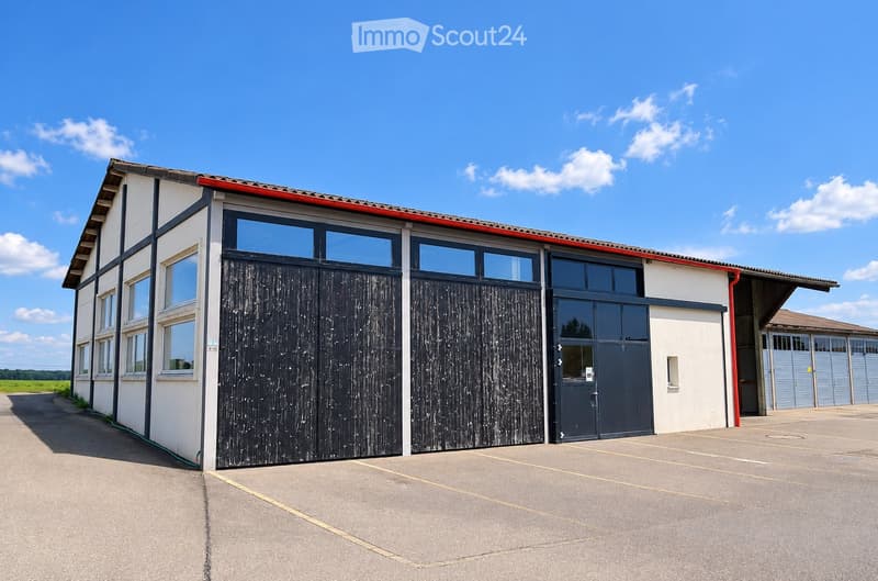 Modern single-story building with white walls, large windows, and dark wooden garage doors. The structure features a red roof trim and a spacious paved parking area.
