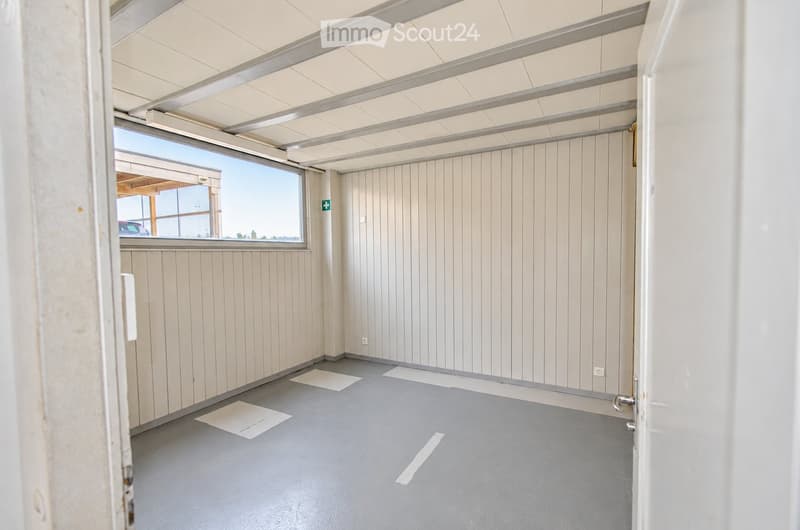 Empty room with light wood panel walls, grey floor, and large windows allowing natural light. No furniture or equipment present.