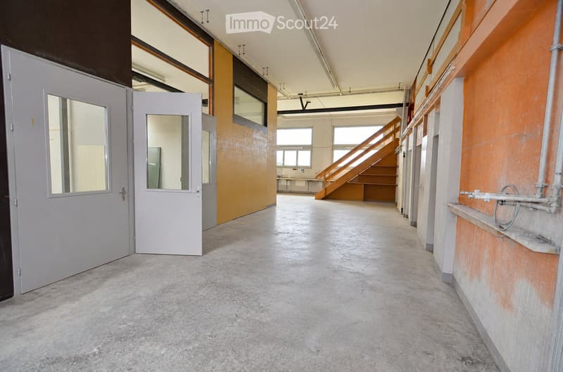 Empty hallway with concrete floor, grey doors, and a staircase leading upwards. Exposed pipes and a small shelf are visible on the right side.