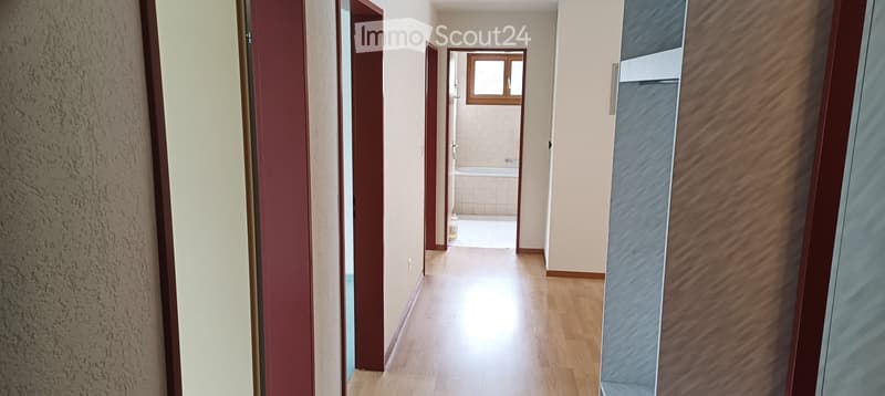 A narrow hallway with wooden flooring, light-colored walls, and several open doors leading to different rooms, including a visible bathroom with a bathtub and tiled walls.