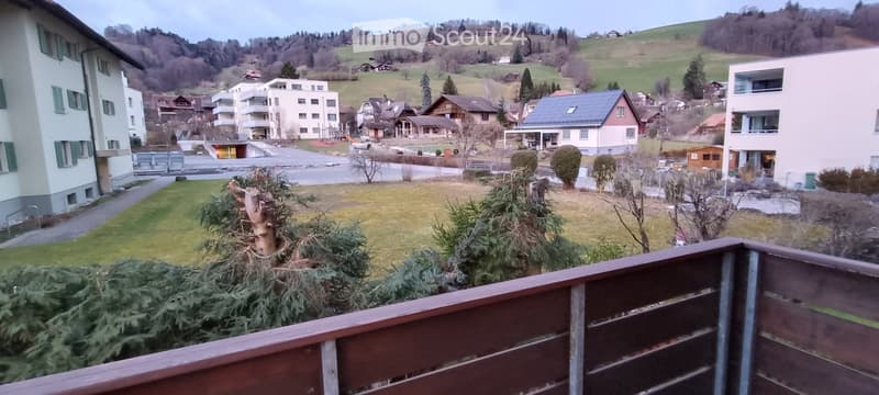 Balcony overlooking a residential area with houses, trees, and a road. The view includes a mix of modern and traditional buildings, green spaces, and a hilly landscape in the background.