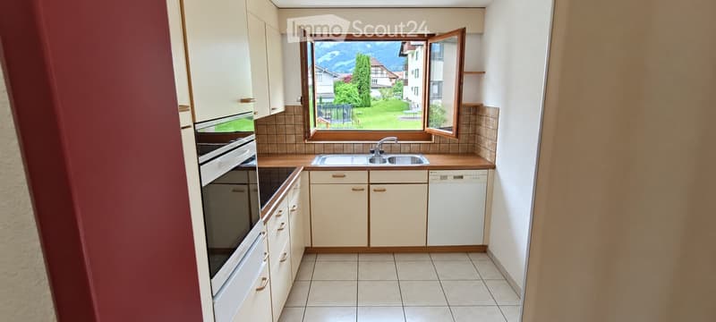 Bright kitchen with white cabinets, a double sink, an oven, and a dishwasher. Large window offers a scenic view of a green landscape and mountains.