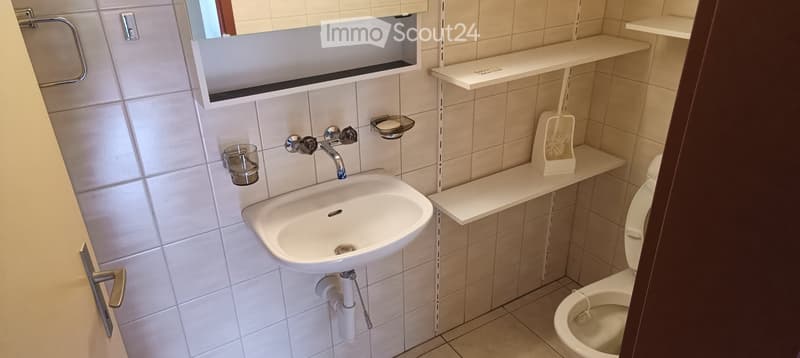 Modern bathroom with white tiled walls, a sink with a chrome faucet, a mirror with built-in shelves, a toilet, and a toilet brush holder on a shelf.