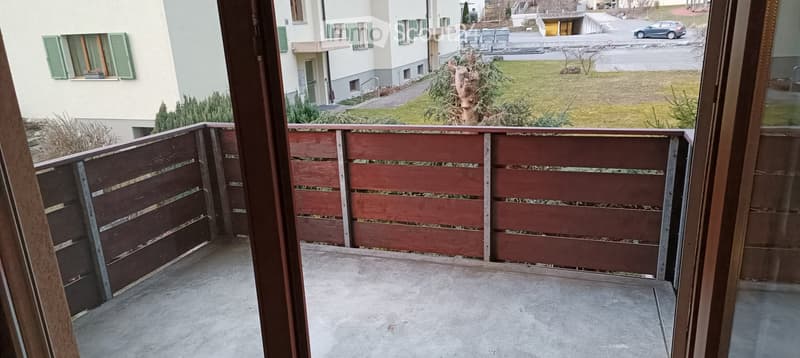 Balcony with wooden railing, concrete floor, overlooking a grassy area and neighboring building.