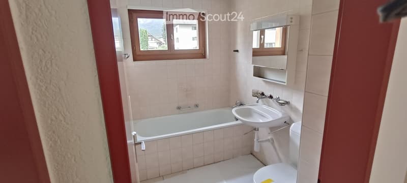 Bathroom with a bathtub, sink, toilet, and mirror. The room has tiled walls and a window with wooden frames.