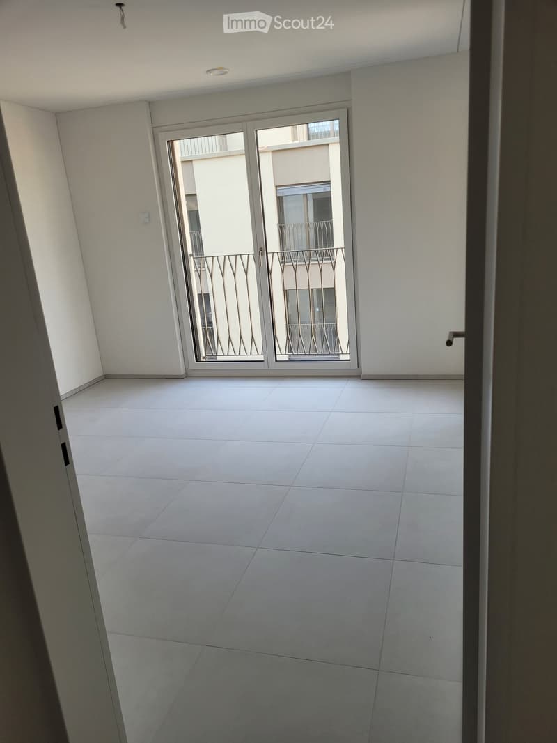 Empty room with white tiled floor and white walls, featuring a large sliding glass door leading to a balcony with metal railings.