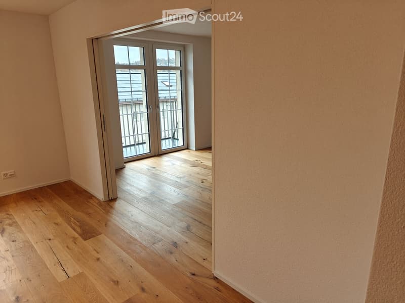 Empty room with light wooden flooring and beige walls, featuring a large sliding glass door leading to a balcony area.