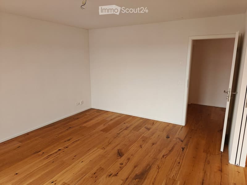 Empty room with light-colored walls and wooden flooring, featuring an open doorway leading to another room.