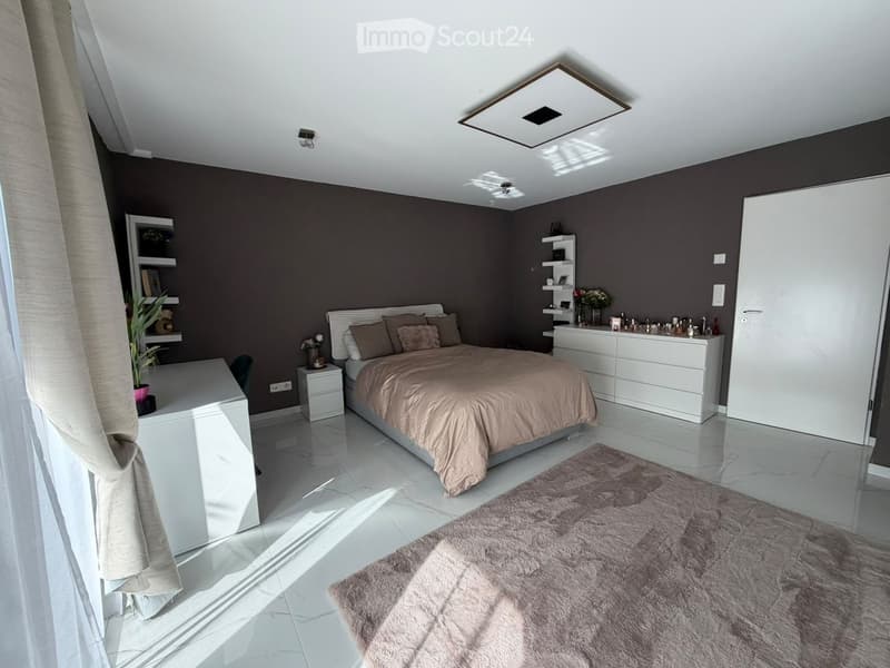 Modern bedroom with a large bed, nightstand, drawer unit, shelves, and a plant. The room features a white tiled floor and dark grey walls.