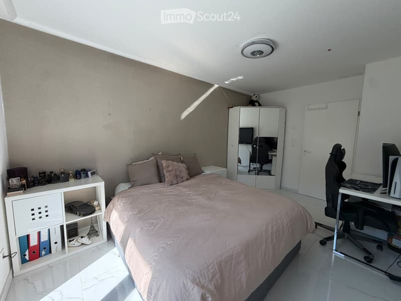 Modern bedroom with a large bed, desk area, and storage cabinet. Features a white shelf with decorative items and a mirror cabinet with a laptop and chair.