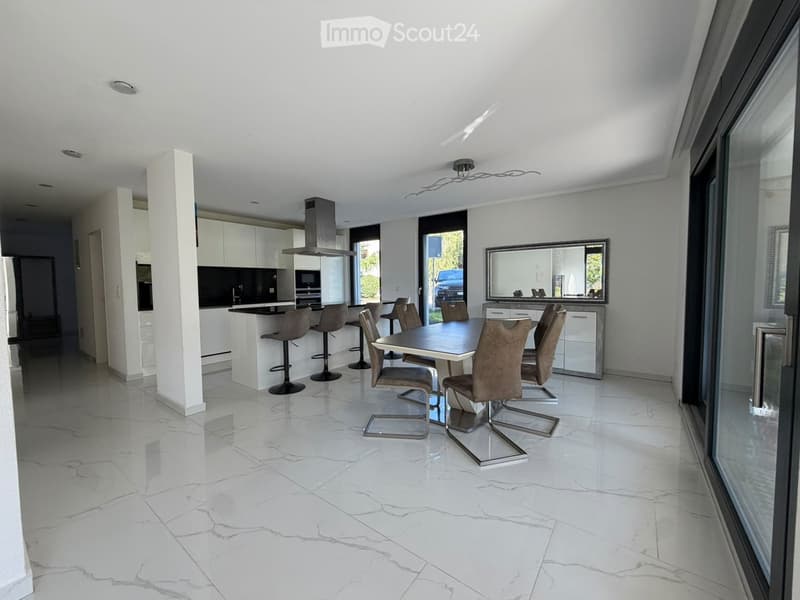 Modern open-plan dining and kitchen area with white marble flooring, sleek white cabinets, a central island with bar stools, and a dining table with chairs.