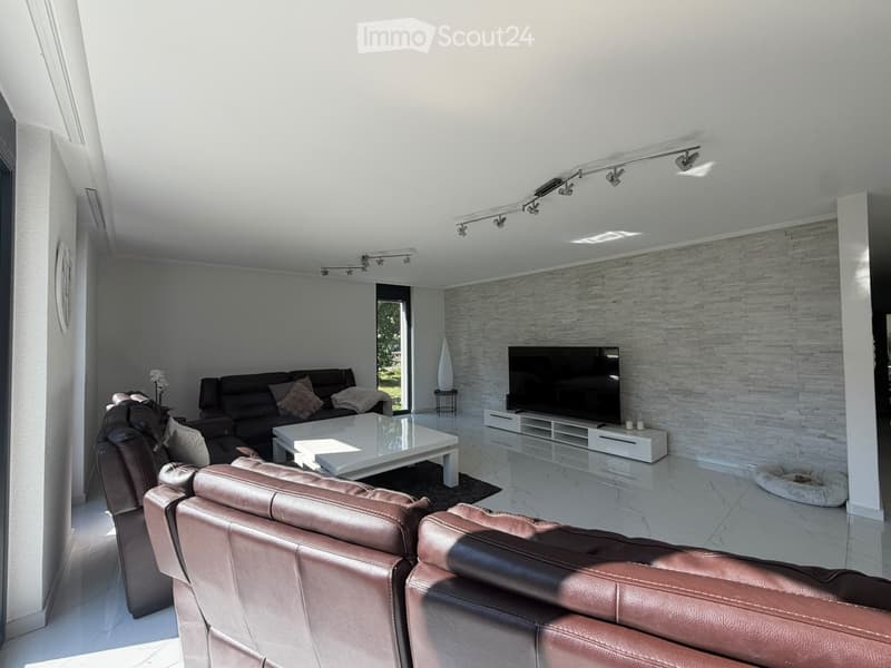 Modern living room with white walls, marble flooring, and a sleek design. Features a black sectional sofa, a white coffee table, a flat-screen TV on a white stand, and additional seating in the foreground.