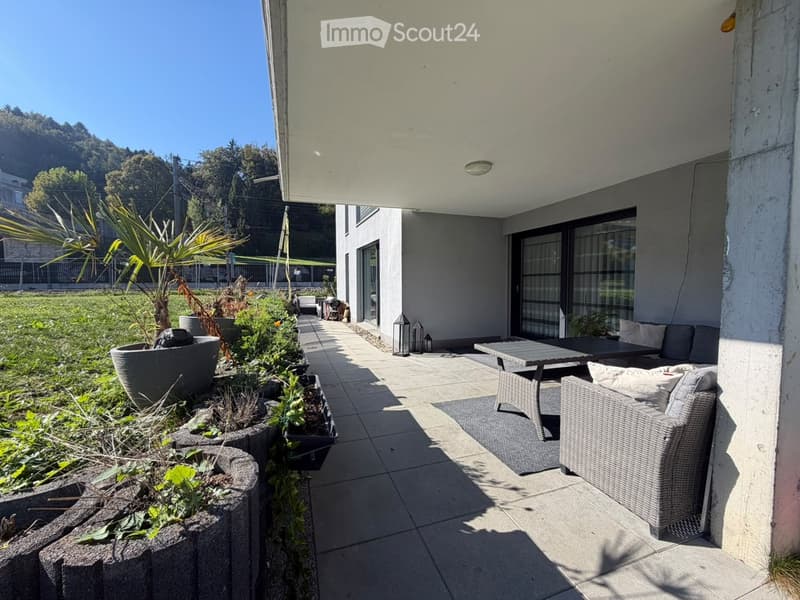 Balcony with a sofa, table, and chairs, surrounded by potted plants and a view of a green lawn.