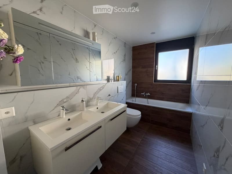 Modern bathroom with double sink, large mirror, bathtub, and toilet. Marble tiles and wooden accents.
