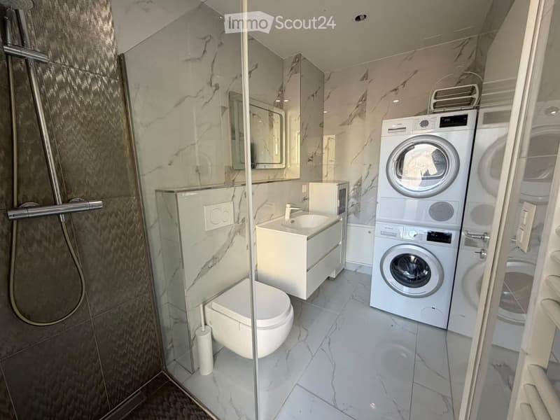 Modern bathroom with marble tiles, a toilet, sink, shower, and stacked washer/dryer unit.
