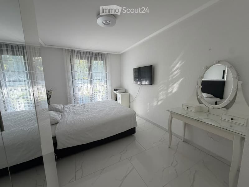 Modern bedroom with a large bed, a vanity table with a mirror, a TV mounted on the wall, and large windows with sheer curtains.