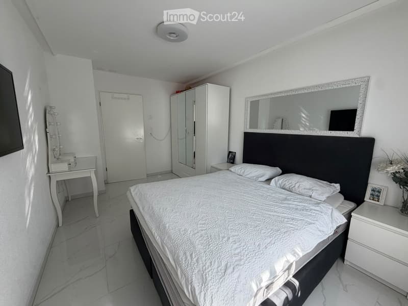 Modern bedroom with a large bed, white bedding, a black headboard, a nightstand with a lamp, a wardrobe, a vanity table, and a large mirror.