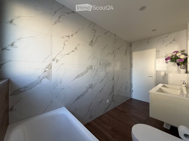 Modern bathroom with marble tiled walls, wooden flooring, bathtub, toilet, sink, and a vase with flowers on the counter.