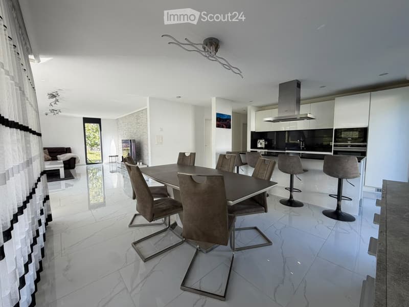 Modern open-plan dining and kitchen area with white cabinets, a dark dining table with grey chairs, and a sofa in the adjacent living space.
