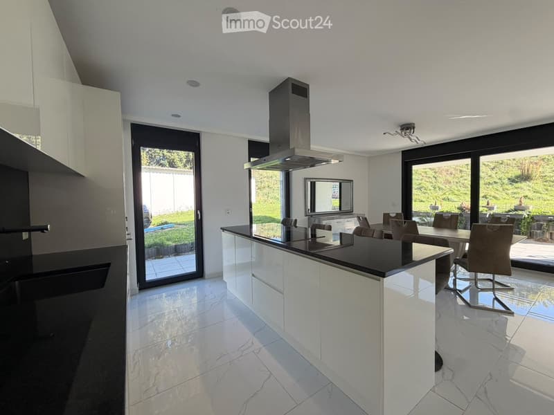 Modern kitchen with white cabinets, black countertops, island, dining area, and large windows overlooking a garden.