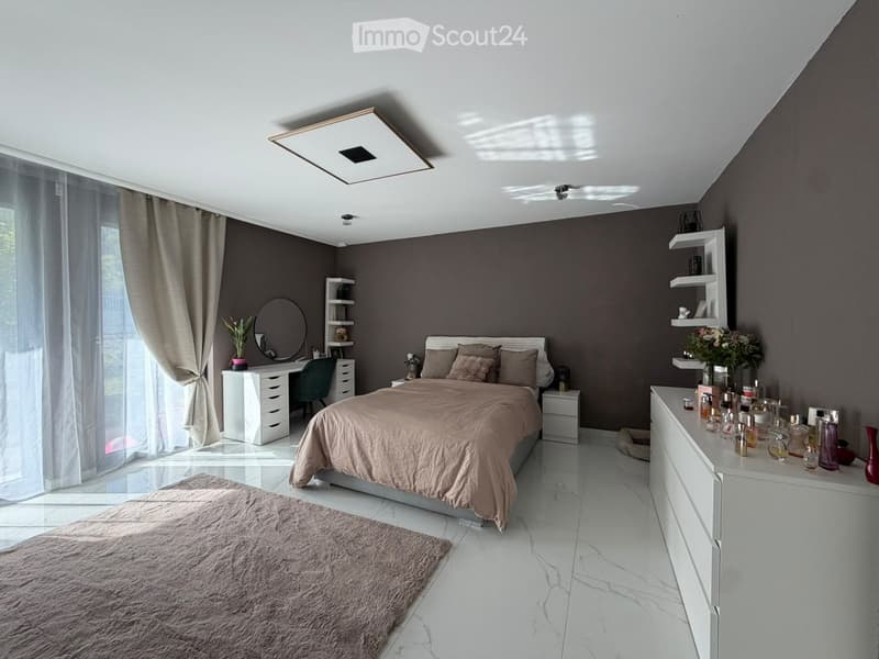 Modern bedroom with a large bed, nightstand, desk, and shelves. Features a sleek design with a gray color palette and natural light from large windows.