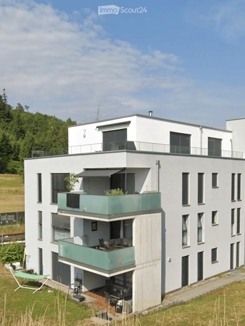 Modern two-story white building with multiple balconies and a rooftop terrace, surrounded by greenery.