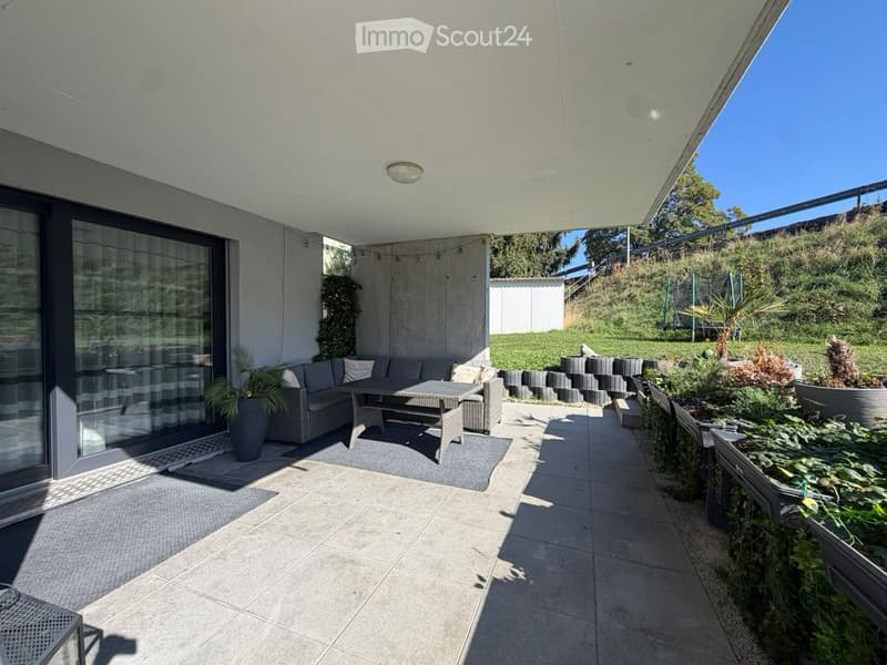 Modern outdoor patio with tiled flooring, grey sofa, coffee table, chairs, and potted plants, overlooking a landscaped garden.