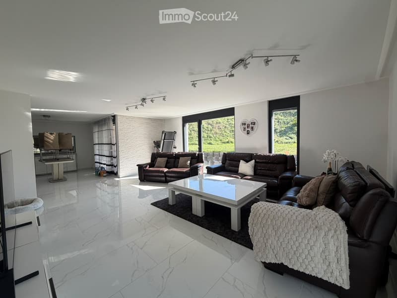 Modern living room with white marble flooring, large windows, and multiple leather sofas arranged around a central coffee table. Features include track lighting, a decorative mirror, and a cozy ottoman with a throw blanket.