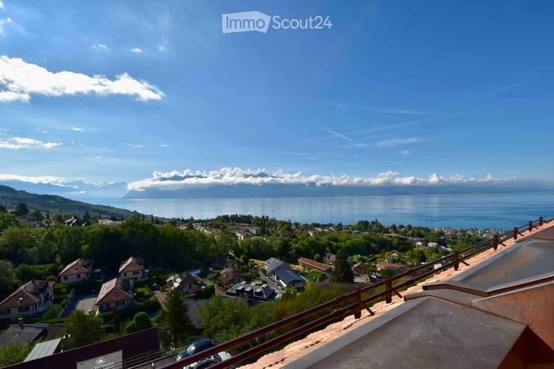 Panoramic view from a rooftop terrace overlooking a town with houses, trees, and a large lake surrounded by mountains under a clear blue sky.