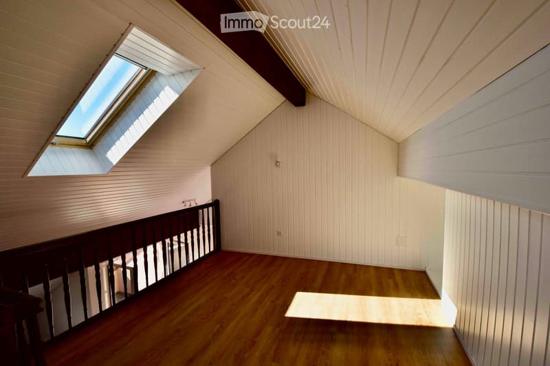 Empty hallway with wooden flooring, white paneled walls, and a skylight window at the top, featuring a dark wooden railing on one side.