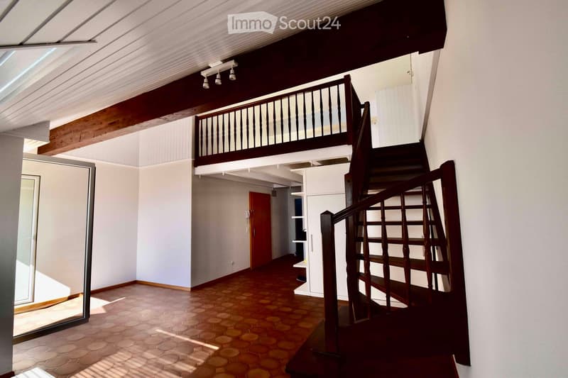 Spacious hallway with wooden staircase leading to an upper level, featuring a wooden railing and a balcony overlooking the lower level. The floor is tiled, and the walls are painted white.