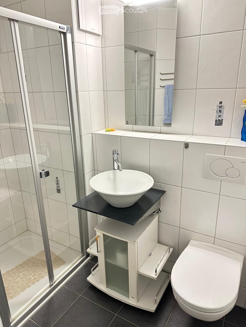 Modern bathroom with white tiles, a glass-enclosed shower, a white sink with a black countertop, a toilet, and a towel hanging on a rack.