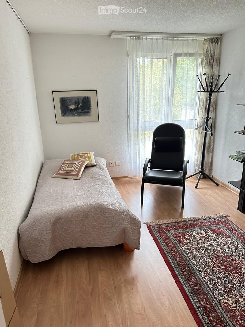 Cozy bedroom with wooden flooring, a single bed with patterned bedding and pillows, a black chair, a coat rack, a picture frame on the wall, and a patterned rug.