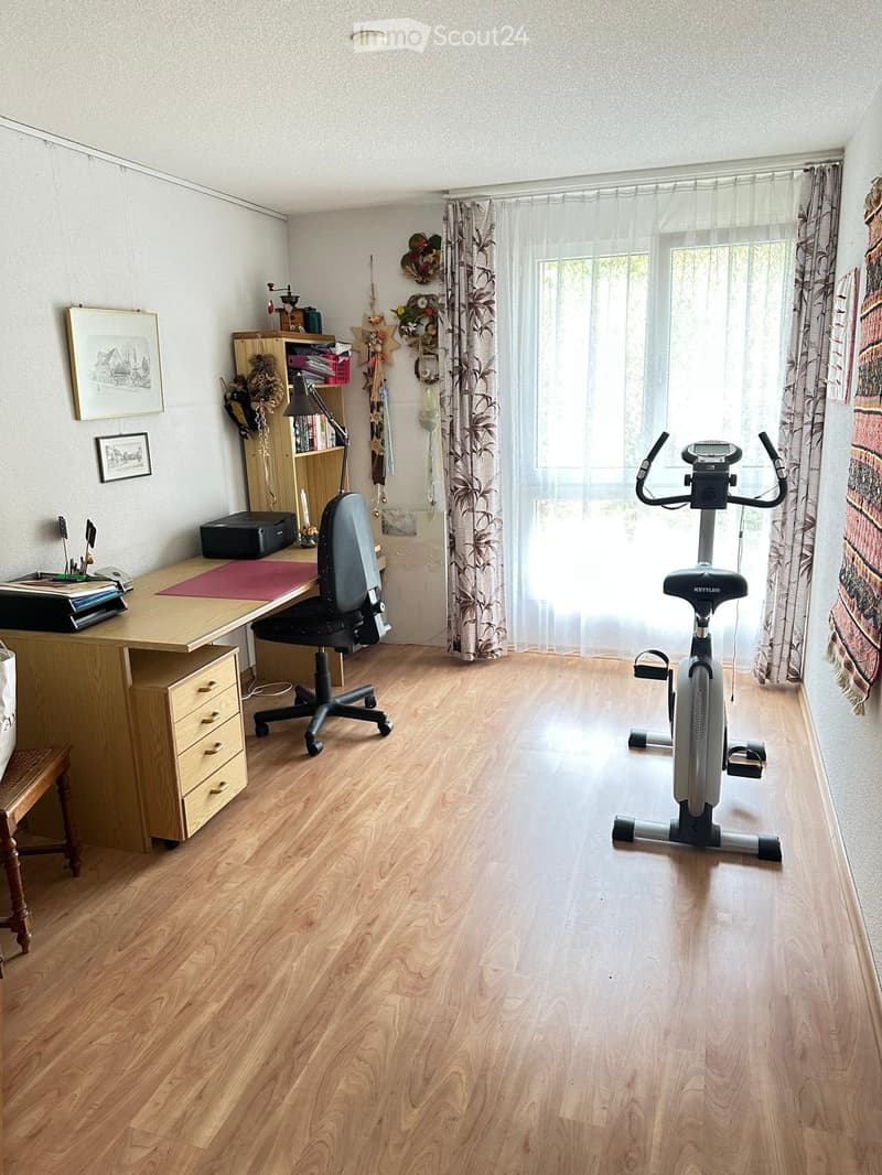 Well-lit room with wooden flooring, a desk with a chair, an exercise bike, a bookshelf, and decorative items on the walls.