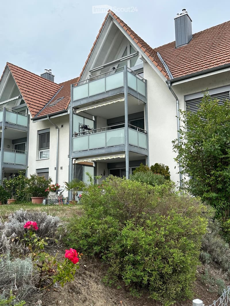 Modern two-story house with red-tiled roof, white walls, and large glass balconies. The house features multiple windows, a chimney, and is surrounded by a well-maintained garden with various plants and flowers.