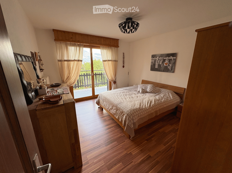 Cozy bedroom with wooden flooring, large bed with checkered bedding, nightstand, dresser with mirror, artwork on wall, and large window with curtains offering a scenic view.