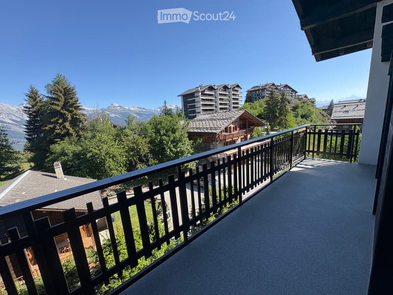 Balcony with a black metal railing, overlooking a scenic mountain view with trees and buildings in the distance.