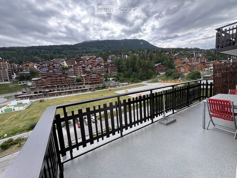 Balcony with a view of a town and mountains, featuring a red chair and a black railing.