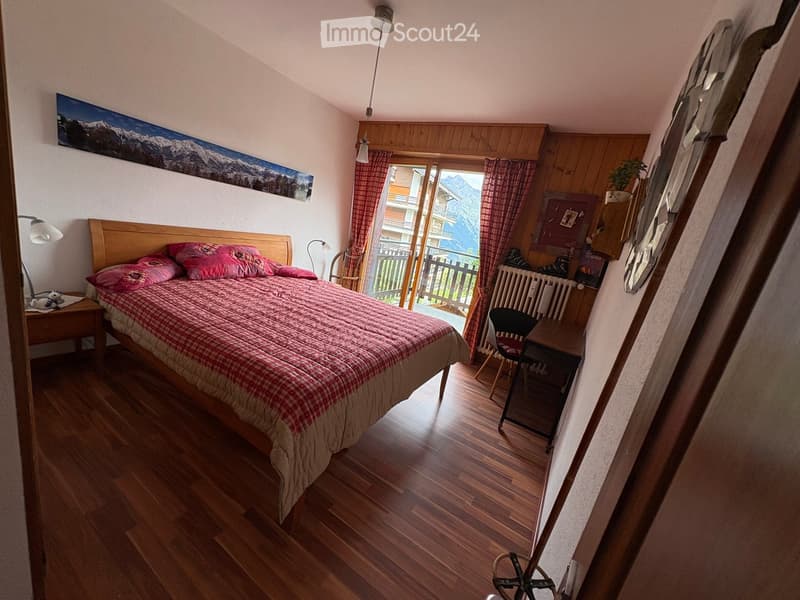 Cozy bedroom with wooden flooring, a large bed with red and white checkered bedding, a nightstand with a lamp, a chair, a small table, and a view of mountains through large windows.