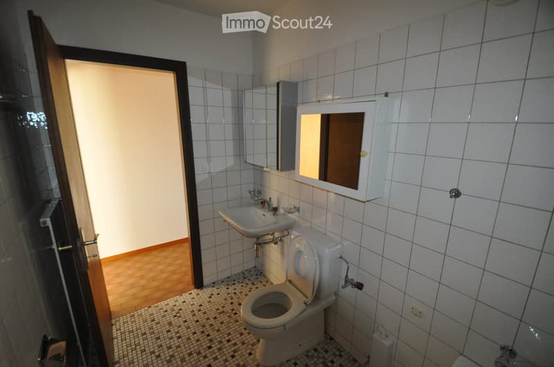 Bathroom with white tiled walls, a toilet, a sink, a mirror with a built-in cabinet, and a wooden door leading to another room.
