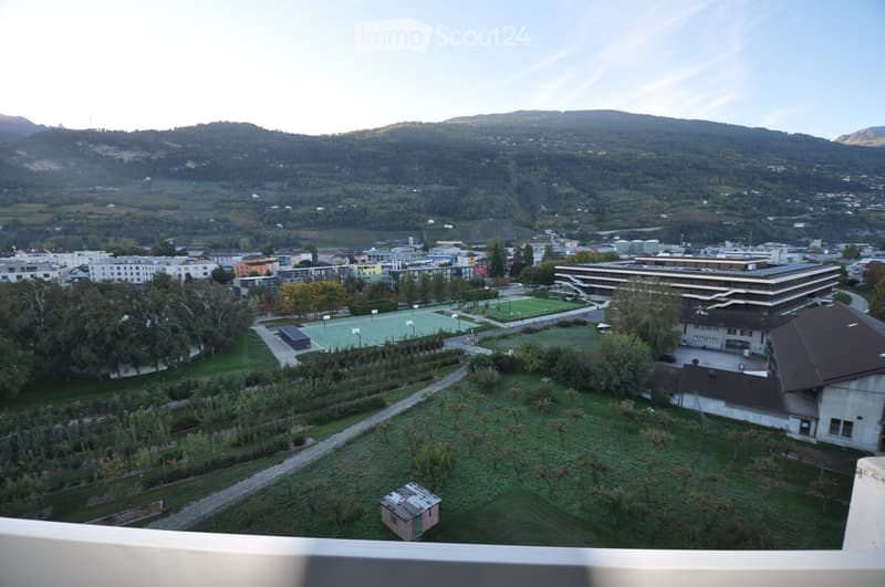 A panoramic view of a suburban area with buildings, green spaces, sports fields, and distant mountains.