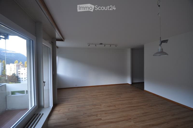Empty room with wooden flooring, white walls, large window with a view, and a hanging light fixture.