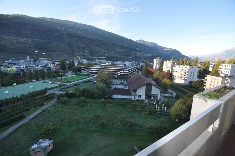 A panoramic view of a town with a green lawn, trees, a small building, and distant mountains in the background.
