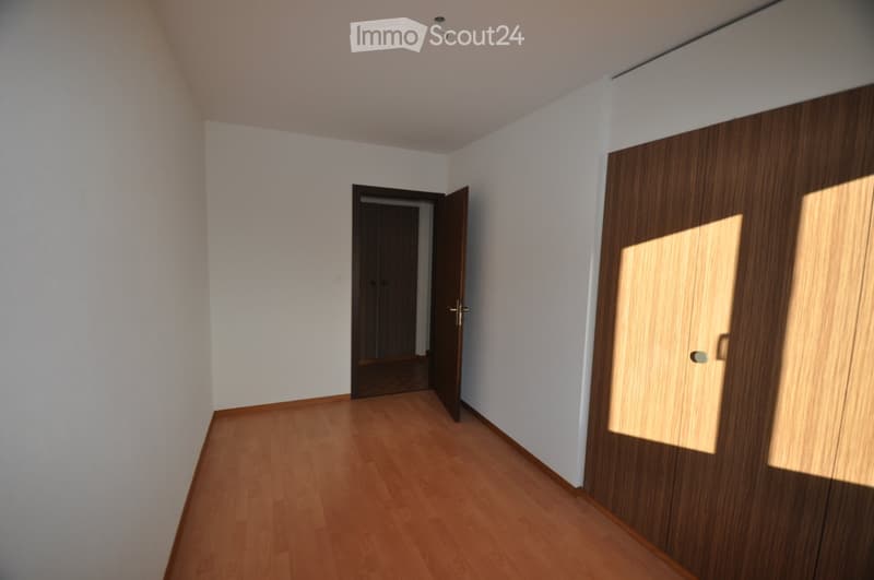 Empty room with wooden flooring, white walls, and a closed wooden wardrobe. An open doorway leads to another room.