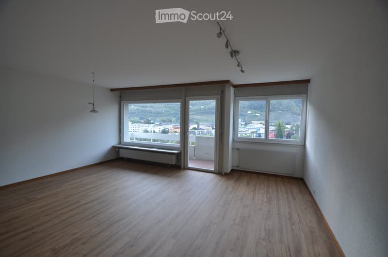 Empty room with wooden flooring, white walls, large windows, and a sliding glass door leading to a balcony.