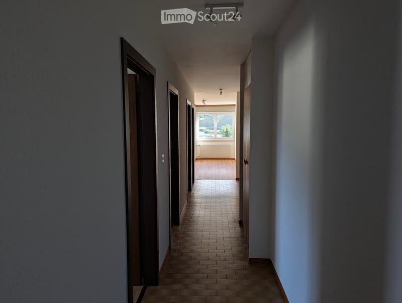 A long, narrow hallway with tiled flooring, wooden doors on both sides, and a view of a room with wooden flooring and large windows at the end.