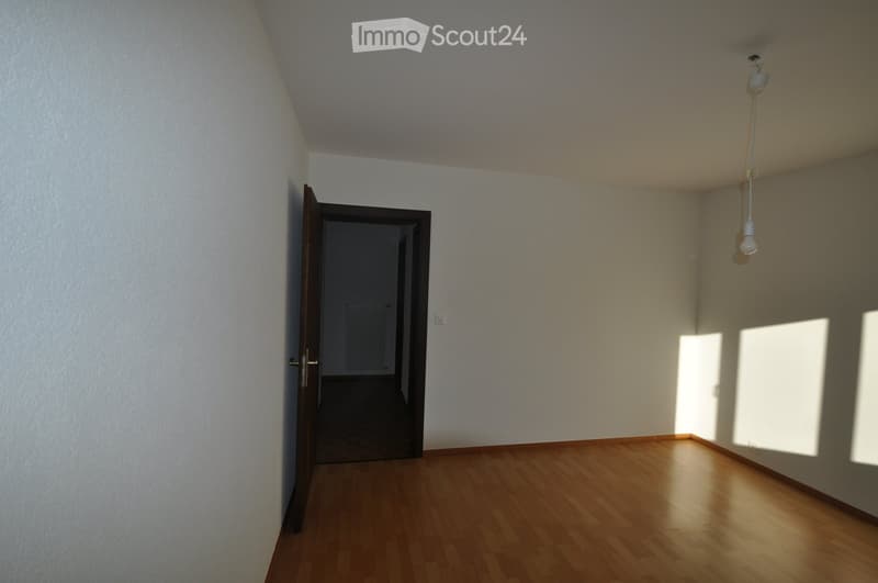 Empty room with wooden flooring, white walls, and a single open door leading to another space. Natural light streams in from the right side.