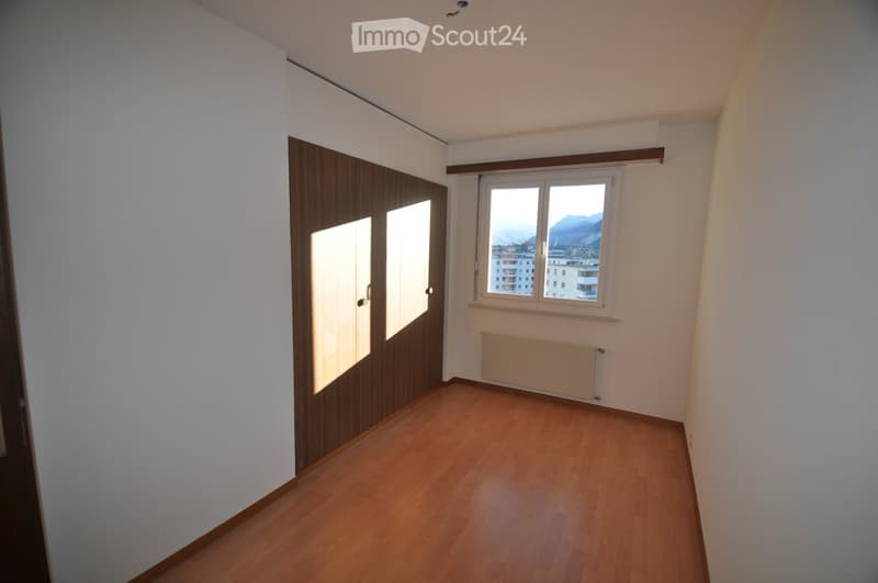 Empty room with wooden flooring, white walls, a large window with a view of mountains and buildings, and a built-in wardrobe with mirrored doors.