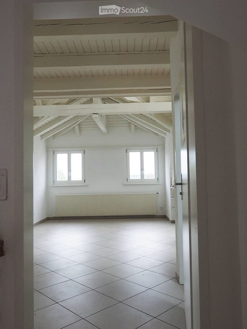 Empty room with white tiled floor, white walls, exposed wooden beams on the ceiling, two windows, and a radiator below one window.