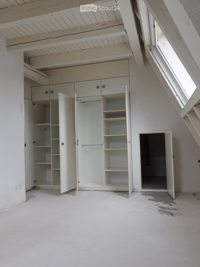 Empty attic room with white walls, exposed wooden beams, built-in white cabinets, and a skylight window. The floor is unfinished concrete with a small water stain.